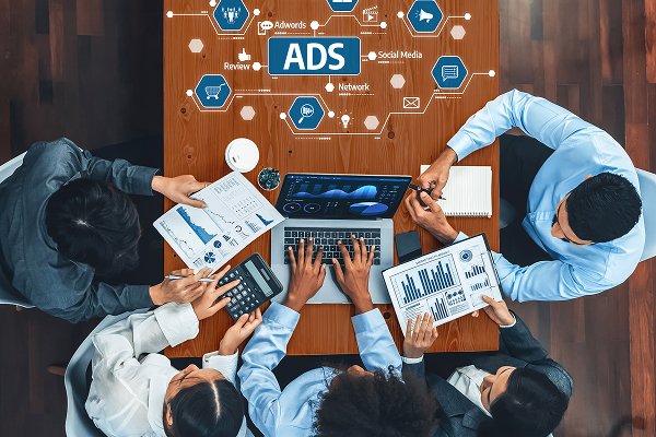 Team collaborating on paid ads management strategy, analyzing data on laptops and documents, featuring "ADS" prominently in digital marketing context.